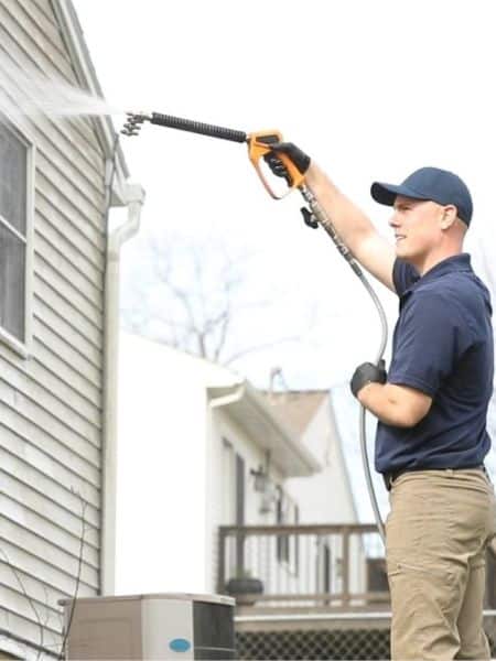 Man power washing the siding of a home with a pressure washer. He wears a blue cap and khakis, aiming the wand upwards. Home maintenance.
