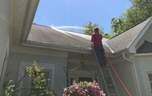 A worker on a ladder uses a power washer to roof clean a residential roof. The man in a red shirt is spraying the asphalt shingles on a sunny day, with trees visible in the background.