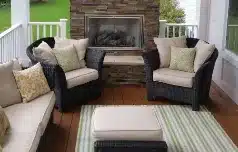 Sunroom with dark wicker sofa and two armchairs with light cushions, striped rug and ottoman with tray, stone fireplace inset in rear wall